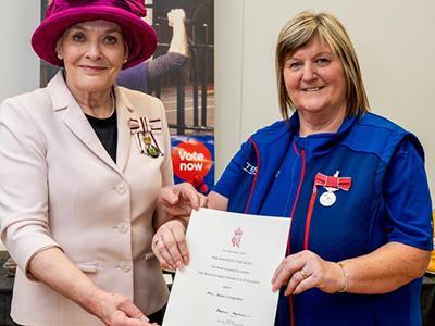 A women presents a certificate to another woman whilst looking at the camera.
