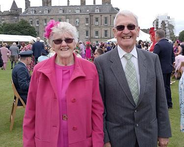 Women in pink suit and a man in a suit stand outside in a garden.