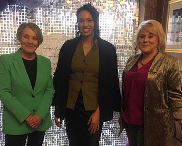 Three women one in a green jacket, one in a black jacket and one in a gold jacket smile at the camera.