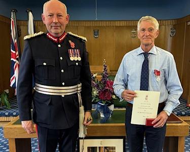 Man in a suit and medals presents a certificate to a man in a suit.