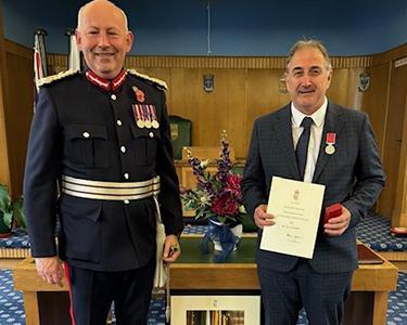 A man in a suit with medals presents a certificate to a man in a suit.