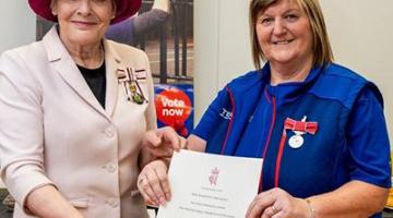 A women presents a certificate to another woman whilst looking at the camera.