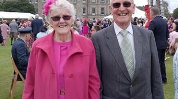 Women in pink suit and a man in a suit stand outside in a garden.