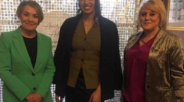 Three women one in a green jacket, one in a black jacket and one in a gold jacket smile at the camera.