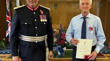 Man in a suit and medals presents a certificate to a man in a suit.